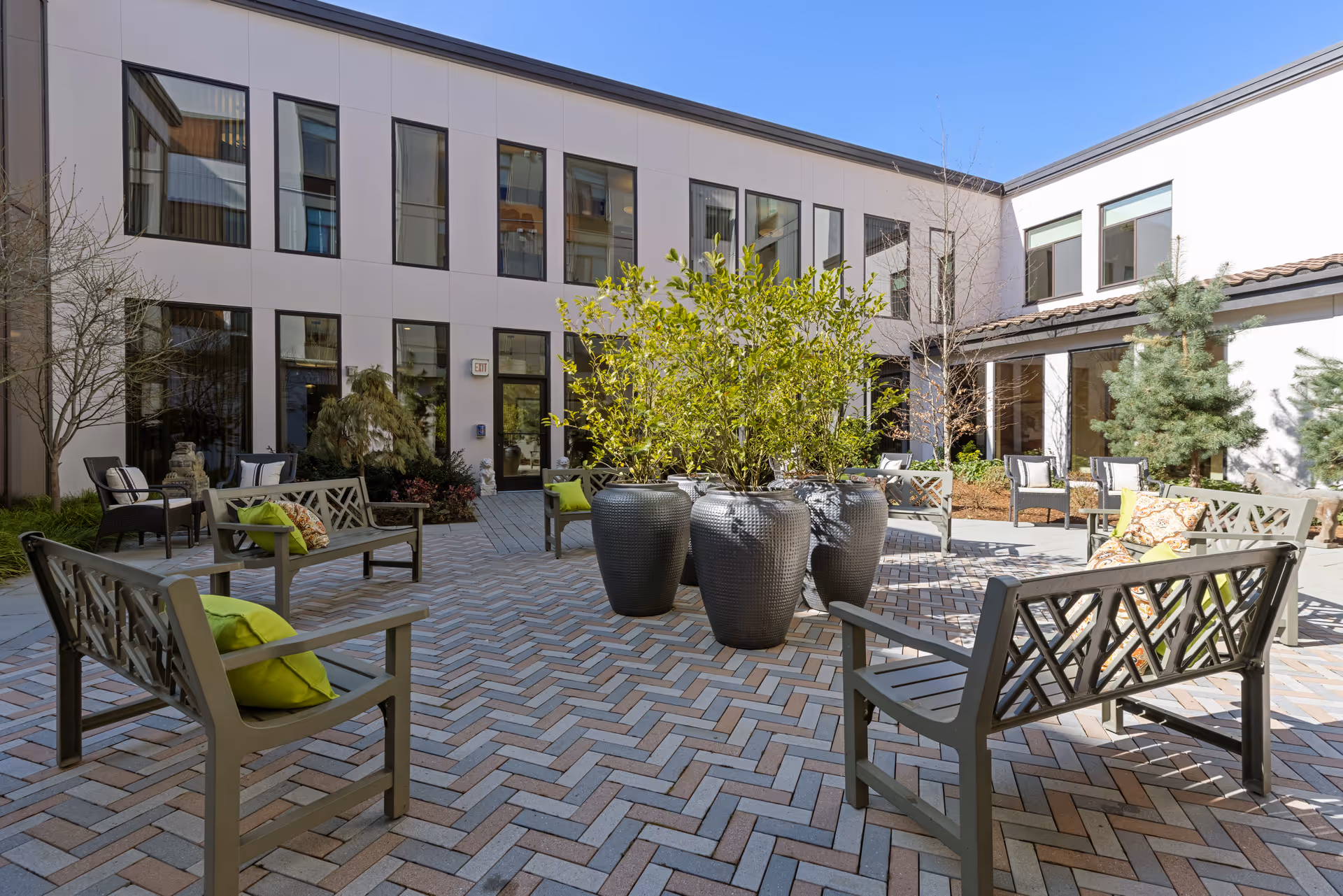 Sunny outdoor courtyard with benches, chairs, large potted plants, and a paved herringbone patio in front of a two-story building.