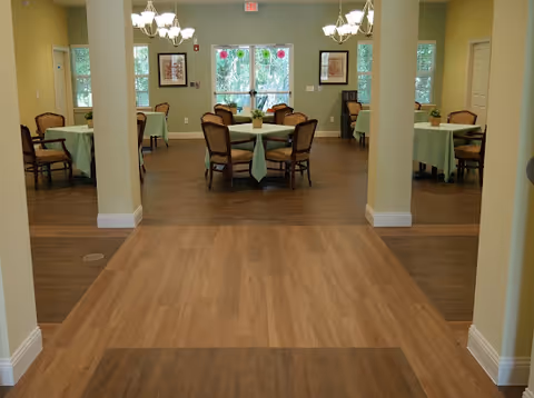 Interior view of a dining room in a senior living facility with several round tables covered with light green tablecloths, each surrounded by four wooden chairs with cushions. The room has light yellow walls, wooden flooring, two chandeliers, framed artwork on the walls, and large windows letting in natural light.