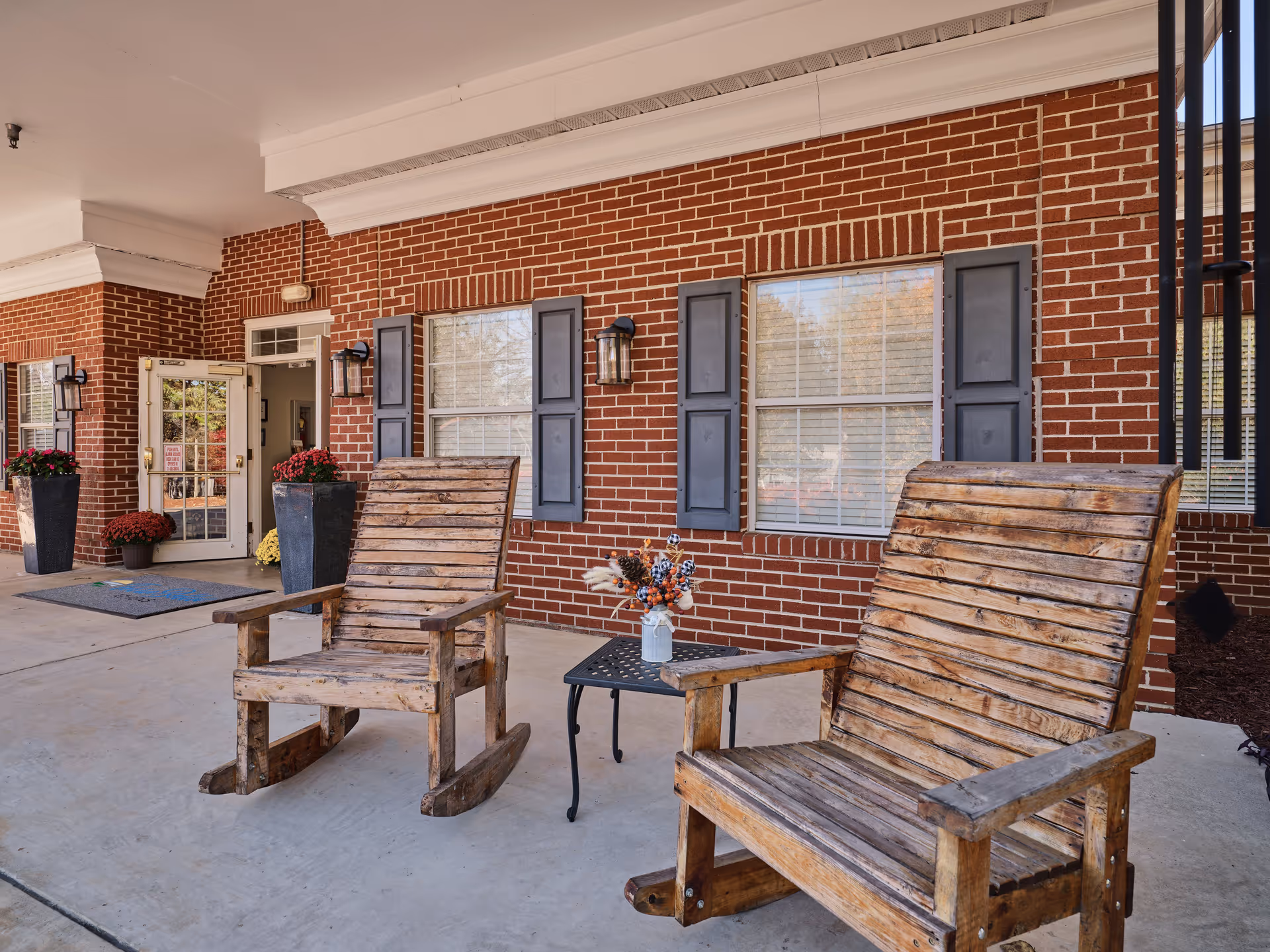Two wooden rocking chairs with a small black metal table holding a vase with flowers between them, placed on a concrete porch in front of a brick building with windows and blue shutters. There are potted plants near the entrance door in the background.