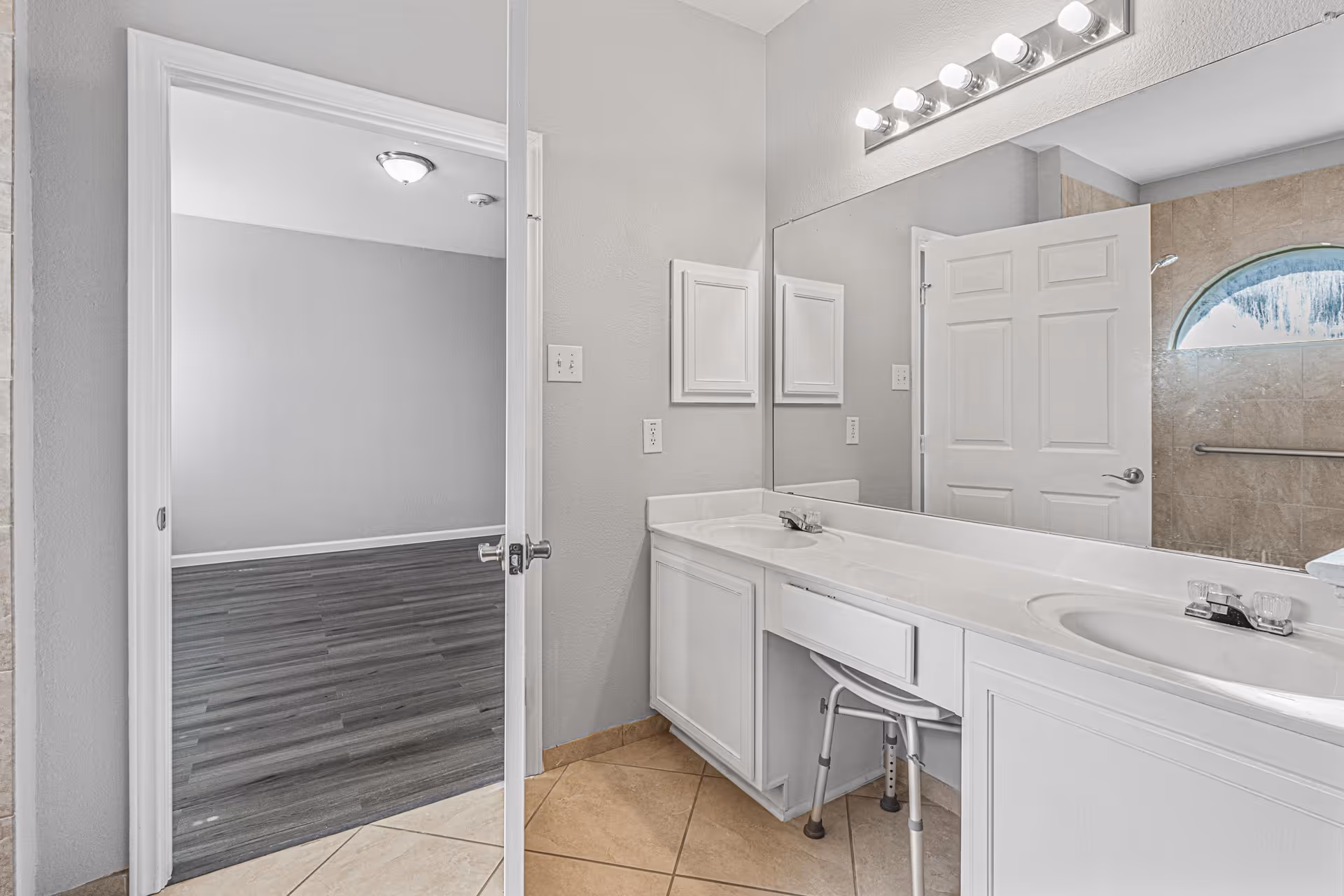 A bathroom with a double sink vanity featuring white cabinets and a large mirror above. There is a small stool under the counter. The bathroom has beige tiled floors and a door leading to a room with dark wood flooring. A shower area with a frosted arched window and a grab bar is visible in the reflection of the mirror.