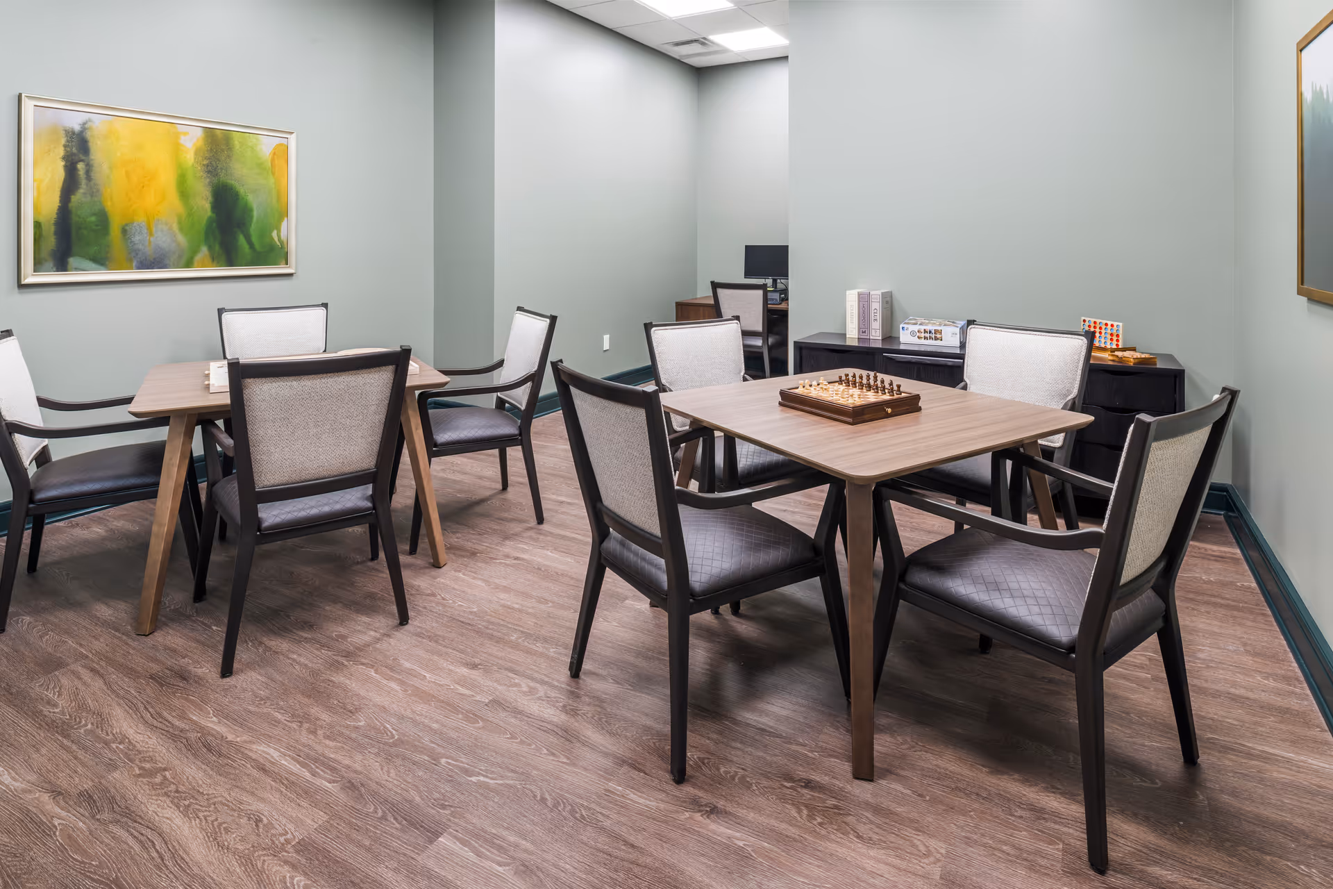 A quiet activity room with two wooden tables, each surrounded by four cushioned chairs. One table has a chessboard set up, and the other has a small game or puzzle. The room has light green walls, wood flooring, and a colorful abstract painting on one wall. In the background, there is a small desk with a computer and some board games on a black cabinet.