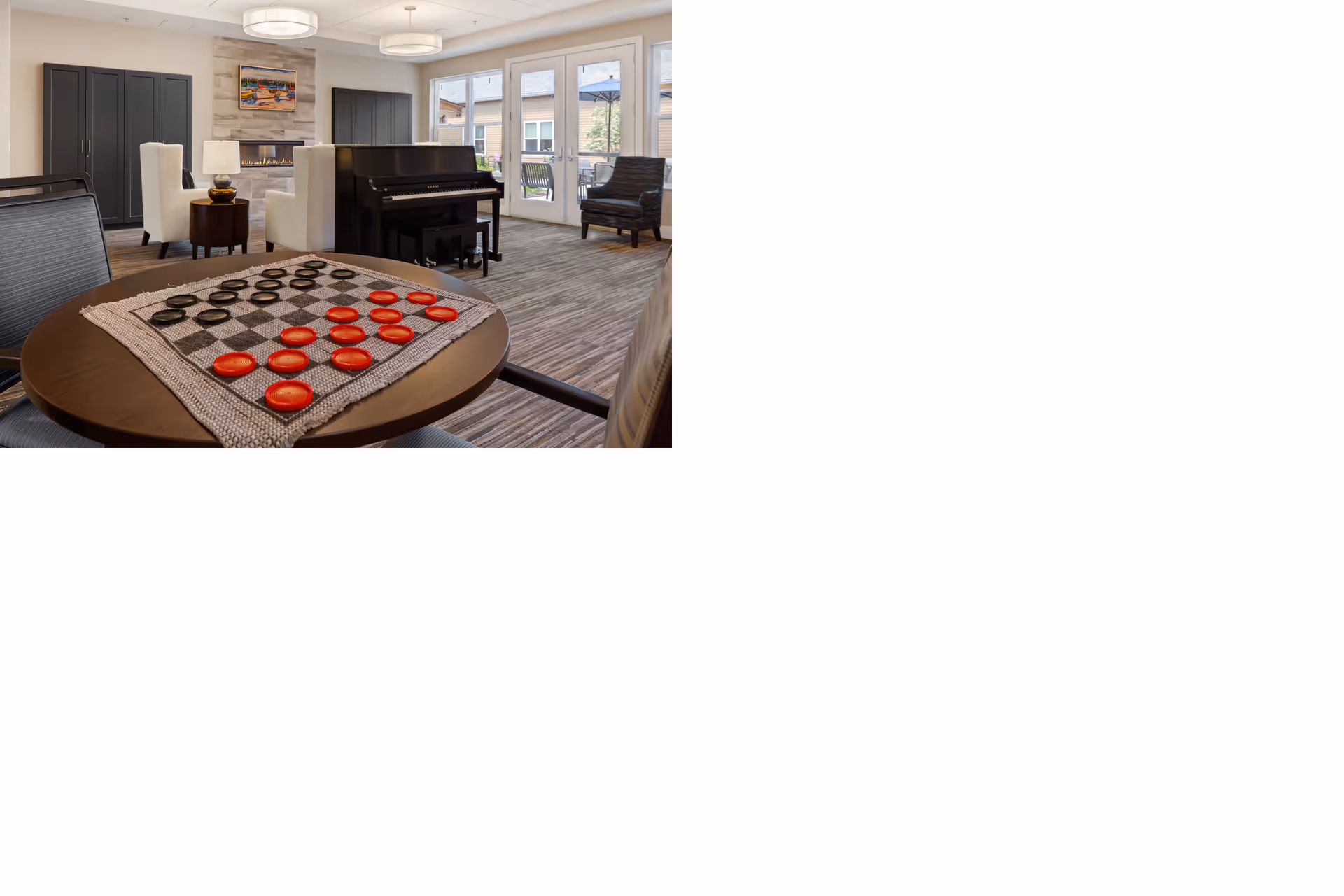 A cozy common area with a round table in the foreground featuring a checkers game set up with red and black pieces. In the background, there is a black piano, white armchairs, a fireplace with a painting above it, and large windows and glass doors leading to an outdoor patio.