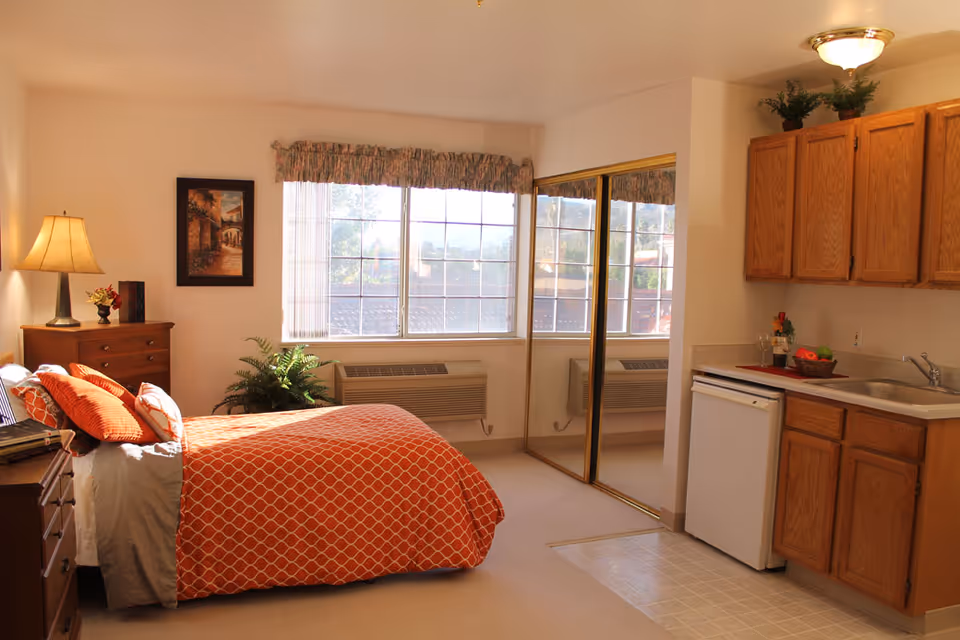 Sunlit studio-style bedroom with an orange-patterned bed, dresser and a small kitchenette with wooden cabinets.