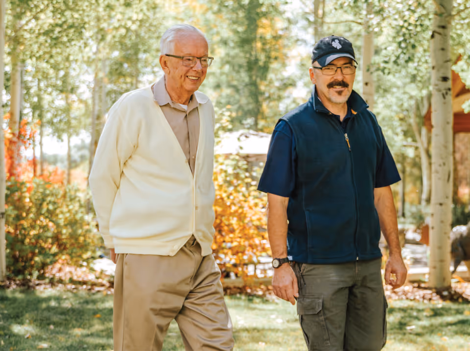 Two men walking outdoors in a garden area with trees and greenery around them. One man is elderly, wearing glasses, a white cardigan, and beige pants. The other man is middle-aged, wearing a black cap, glasses, a navy blue vest, and cargo pants. They appear to be enjoying a sunny day.