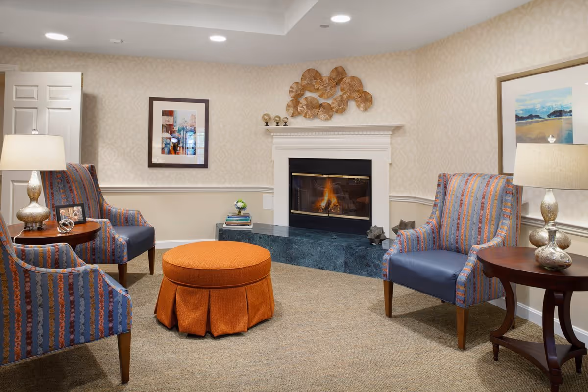 A cozy living room with a lit fireplace, two patterned armchairs with blue cushions, an orange round ottoman, two wooden side tables with lamps, framed artwork on the walls, and decorative items on the fireplace mantel and side table.