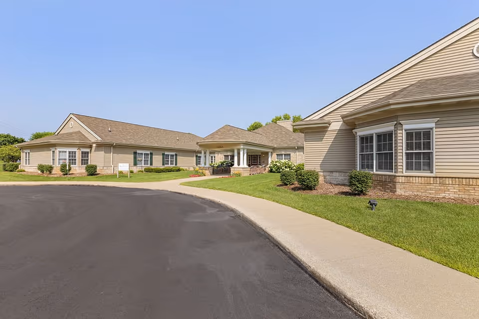 Exterior view of a single-story senior living facility building with beige siding and a covered entrance. The building is surrounded by a well-maintained lawn, shrubs, and a paved driveway under a clear blue sky.