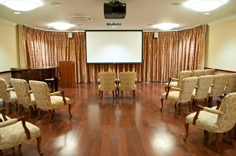 A small auditorium or meeting room with wooden flooring, upholstered armchairs arranged in rows facing a white projection screen, a wooden podium, and a black grand piano in the corner. The back wall is covered with patterned curtains.