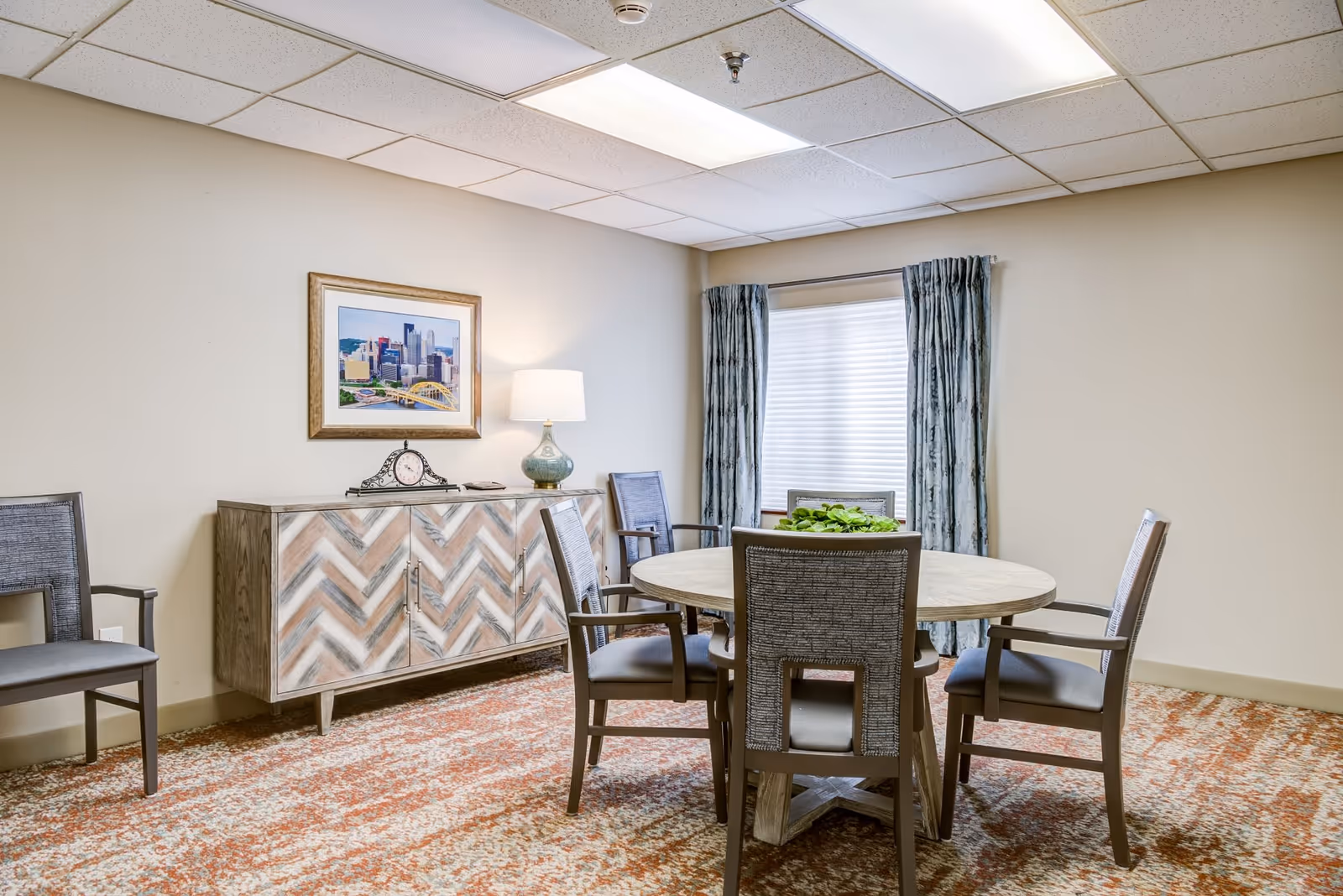 A small dining area with a round wooden table surrounded by five gray cushioned chairs. A sideboard with a chevron pattern stands against the wall, topped with a decorative clock and a lamp. A framed picture of a cityscape hangs above the sideboard. The room has beige walls, a window with blue curtains, and a patterned carpet in shades of orange and beige.