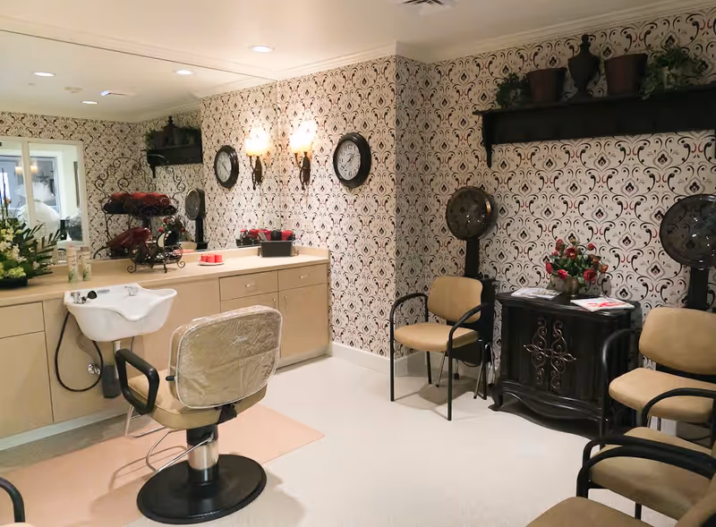 Interior of a hair salon area in a senior living facility with a salon chair in front of a sink and counter, patterned wallpaper, two wall clocks, two hair dryers, beige chairs, a small cabinet with flowers and magazines, and decorative wall sconces.
