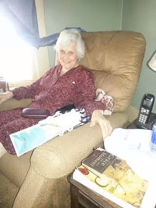 An elderly woman with white hair wearing a patterned maroon outfit is sitting comfortably in a beige recliner chair in a room with green walls. There is a window with a curtain behind her, a cordless phone on a small table next to the chair, and two books on the table. The woman is smiling and holding a light blue booklet.