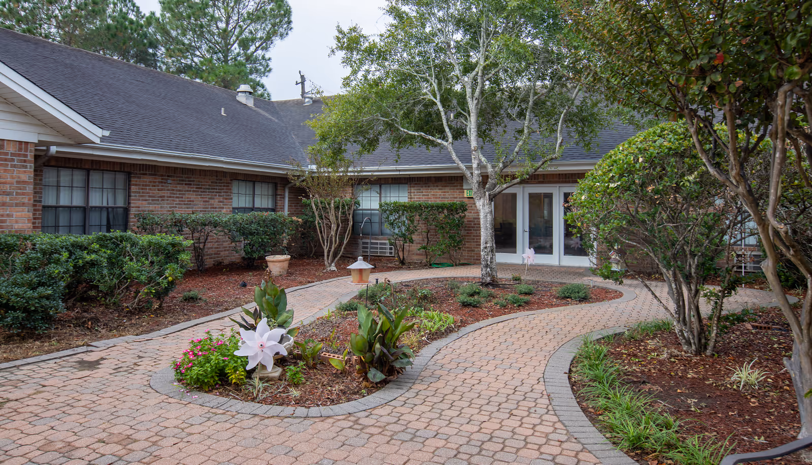 A landscaped courtyard with a brick-paved walkway winding through garden beds containing shrubs, small plants, and a tree. The courtyard is surrounded by a single-story brick building with windows and white double doors.
