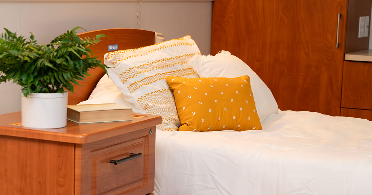 A neatly made bed with white and yellow pillows in a room with wooden furniture, including a nightstand with a potted fern and a closed book on it.