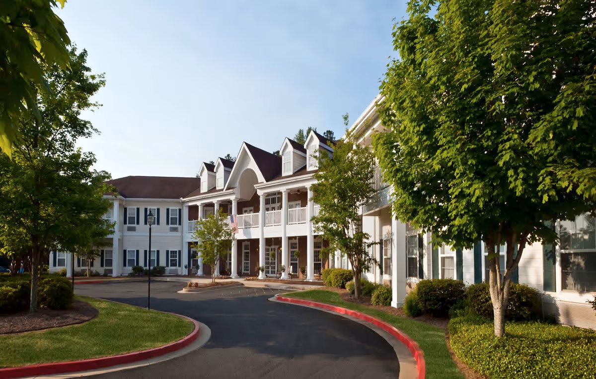 Exterior view of a two-story senior living facility building with white siding, multiple windows, and a covered entrance with columns. There is a curved driveway in front, surrounded by well-maintained landscaping including green trees and bushes under a clear blue sky.