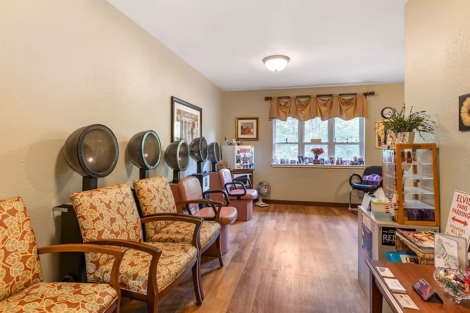 Small salon-style room with several hooded hair dryers and upholstered chairs along one wall, a window at the far end, and a table with brochures on the right.