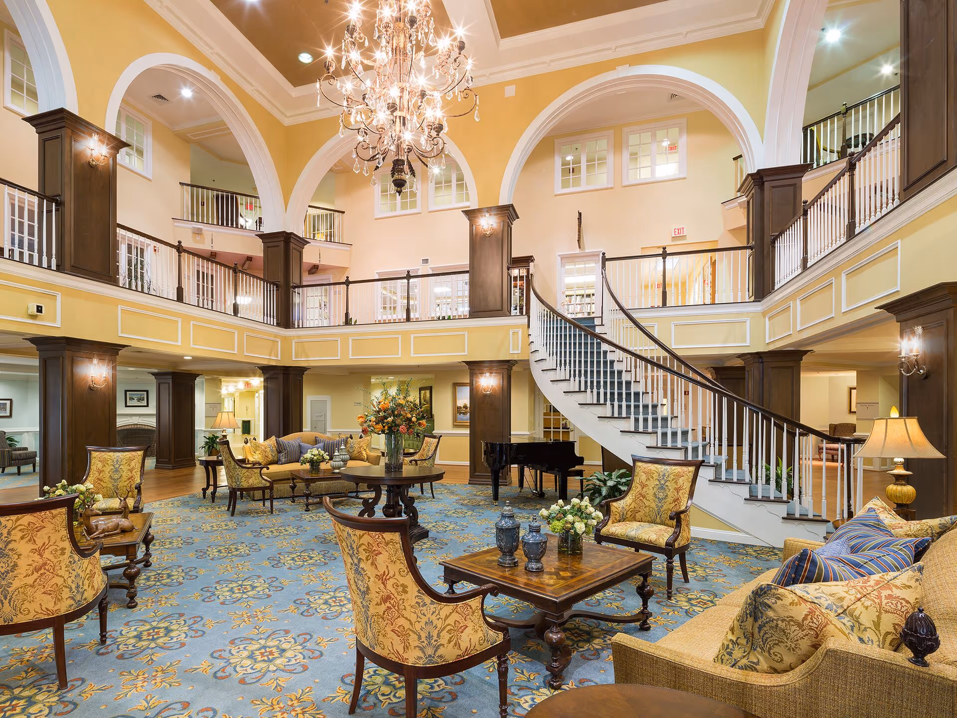 A spacious and elegant living room area in a senior living facility with high ceilings, large chandelier, a curved staircase, and a balcony overlooking the room. The room is furnished with patterned armchairs, sofas with decorative pillows, wooden coffee tables, and floral arrangements. The walls are painted yellow with white trim and dark wood columns.