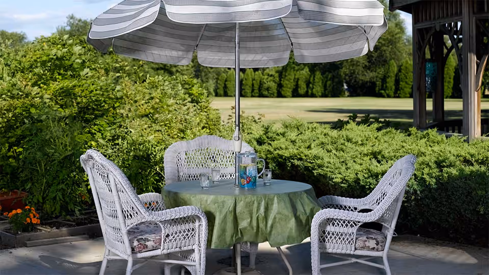 Outdoor patio area with a round table covered by a green tablecloth, surrounded by four white wicker chairs with cushions. A large striped umbrella shades the table, which has a pitcher of water and glasses on it. The background features green bushes, trees, and a grassy field under a clear sky.