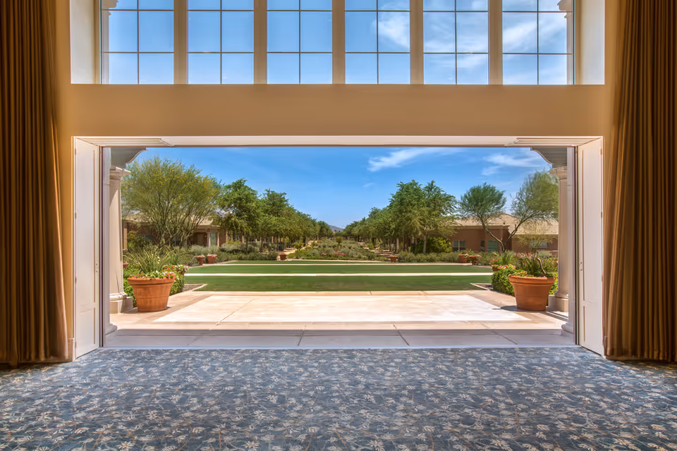 View from inside a building looking out through a large open doorway onto a landscaped garden area with green grass, trees, and potted plants under a blue sky with some clouds. The interior has patterned carpet and tall windows above the doorway.