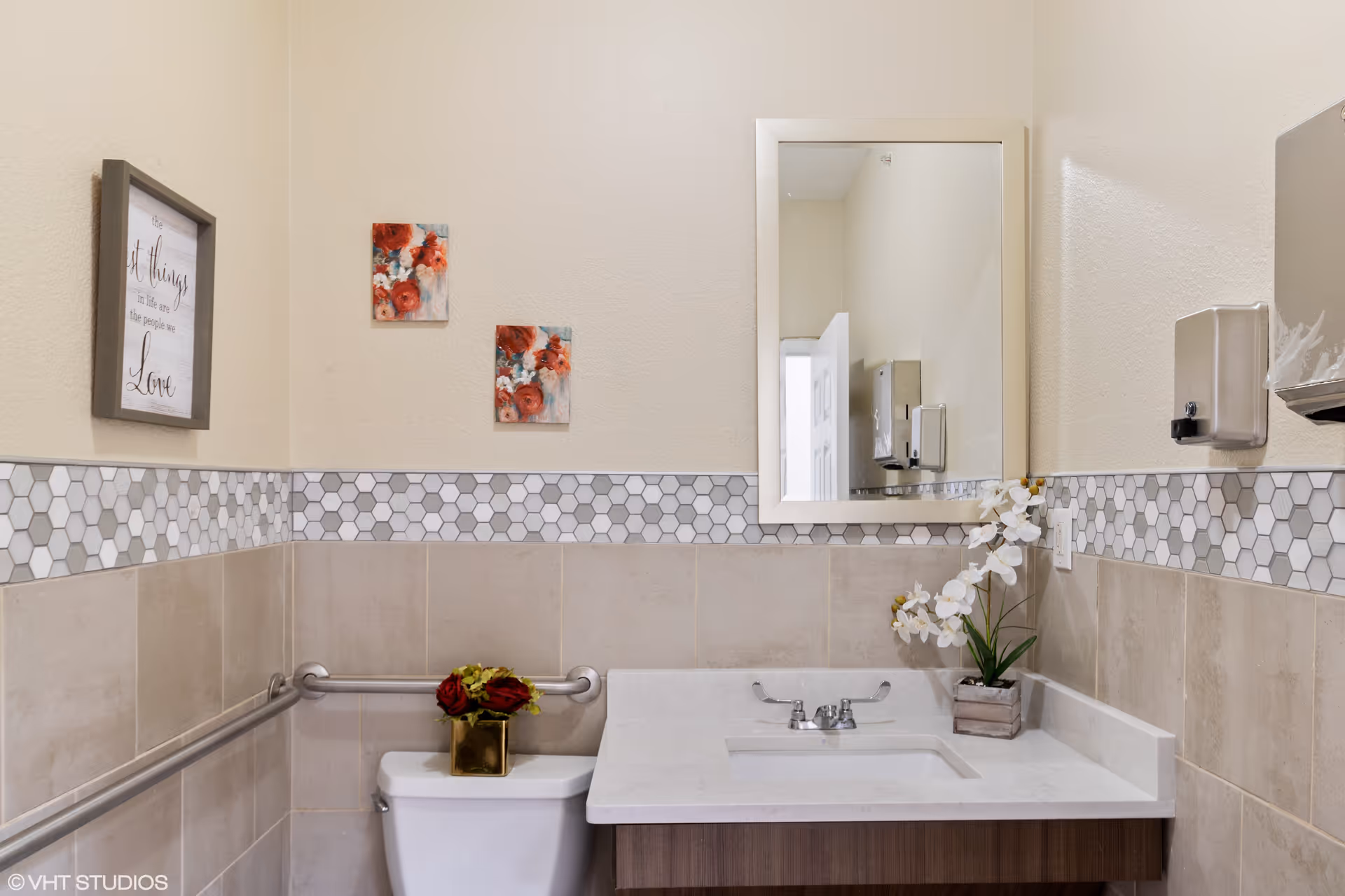A clean bathroom with beige tiled walls and a white countertop sink. There is a rectangular mirror above the sink, a white toilet with a small vase of red flowers on top, and a metal handrail on the wall. The walls have a decorative hexagonal tile border and are adorned with two small floral paintings and a framed quote. A soap dispenser is mounted on the wall next to the mirror.