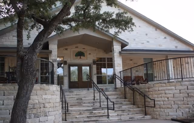 Entrance of a building with stone walls and steps leading up to double wooden doors under a covered porch. There is a tree on the left side and metal railings along the stairs and porch.