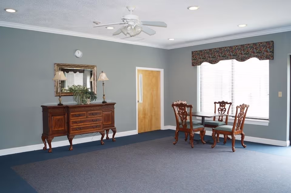 A room with gray walls and blue carpet featuring a wooden sideboard with two lamps and a plant on top, a wall clock above it, a mirror reflecting window blinds, a wooden door, and a small dining table with four ornate wooden chairs near a large window with blinds and a floral valance.