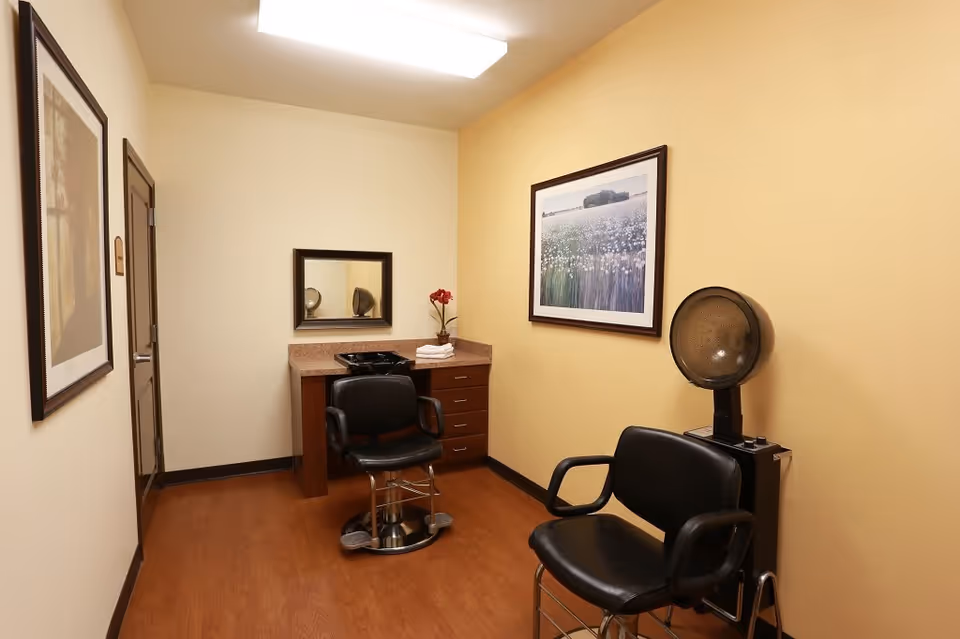 Small salon room with two black styling chairs, a hooded hair dryer, a sink counter and framed artwork on the walls.