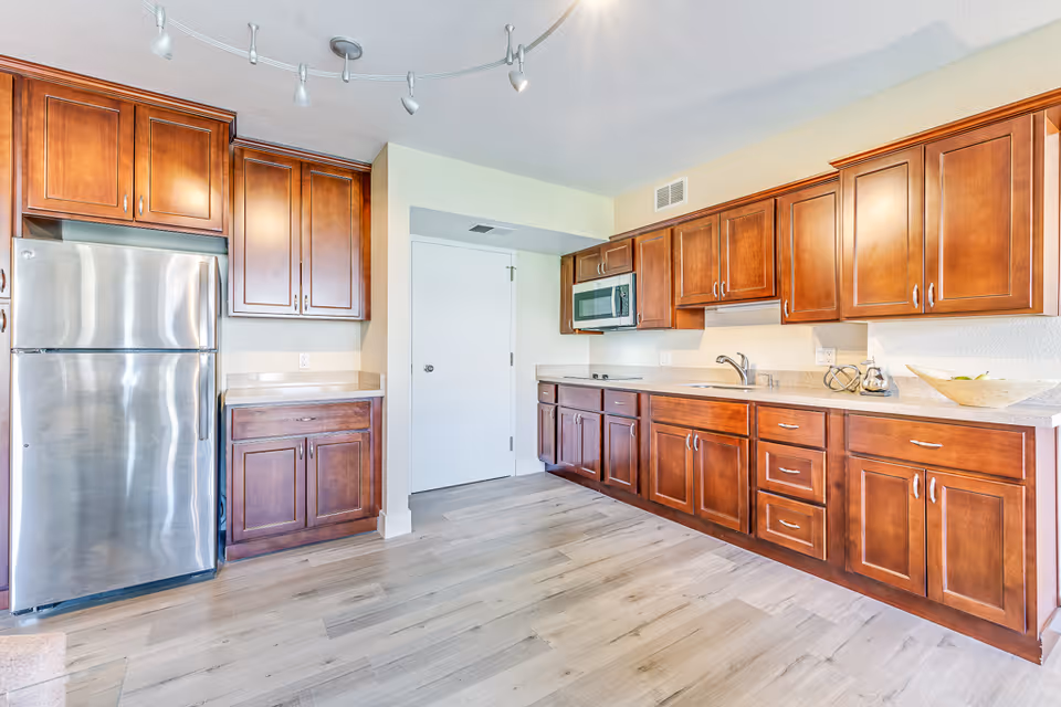 Bright kitchen with wooden cabinets, a stainless steel refrigerator, a built-in microwave, a sink, and light-colored countertops. The floor is light wood, and there is a white door in the background.