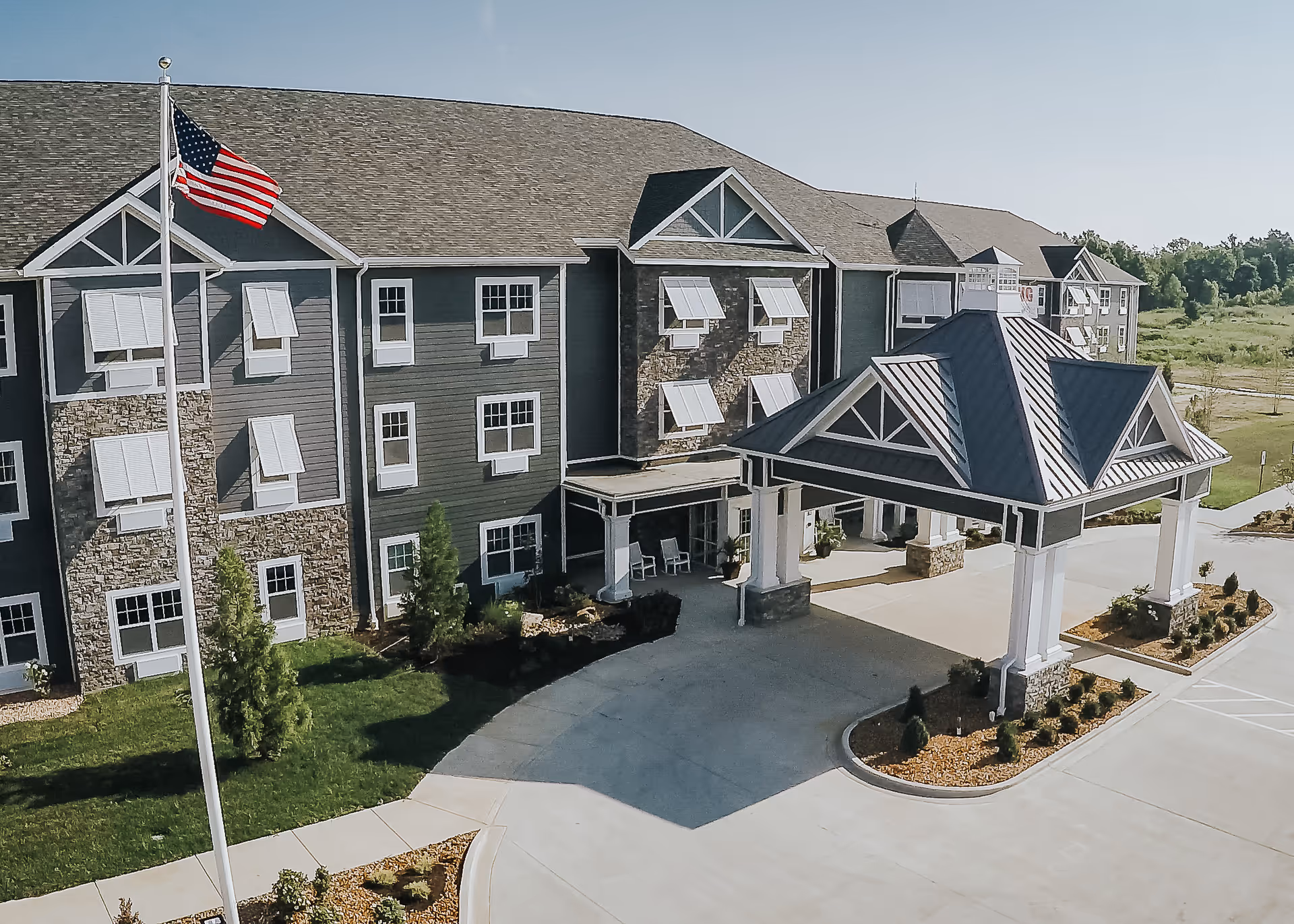 Front exterior of a multi-story senior living building with a covered porte-cochere, landscaping, and an American flag.
