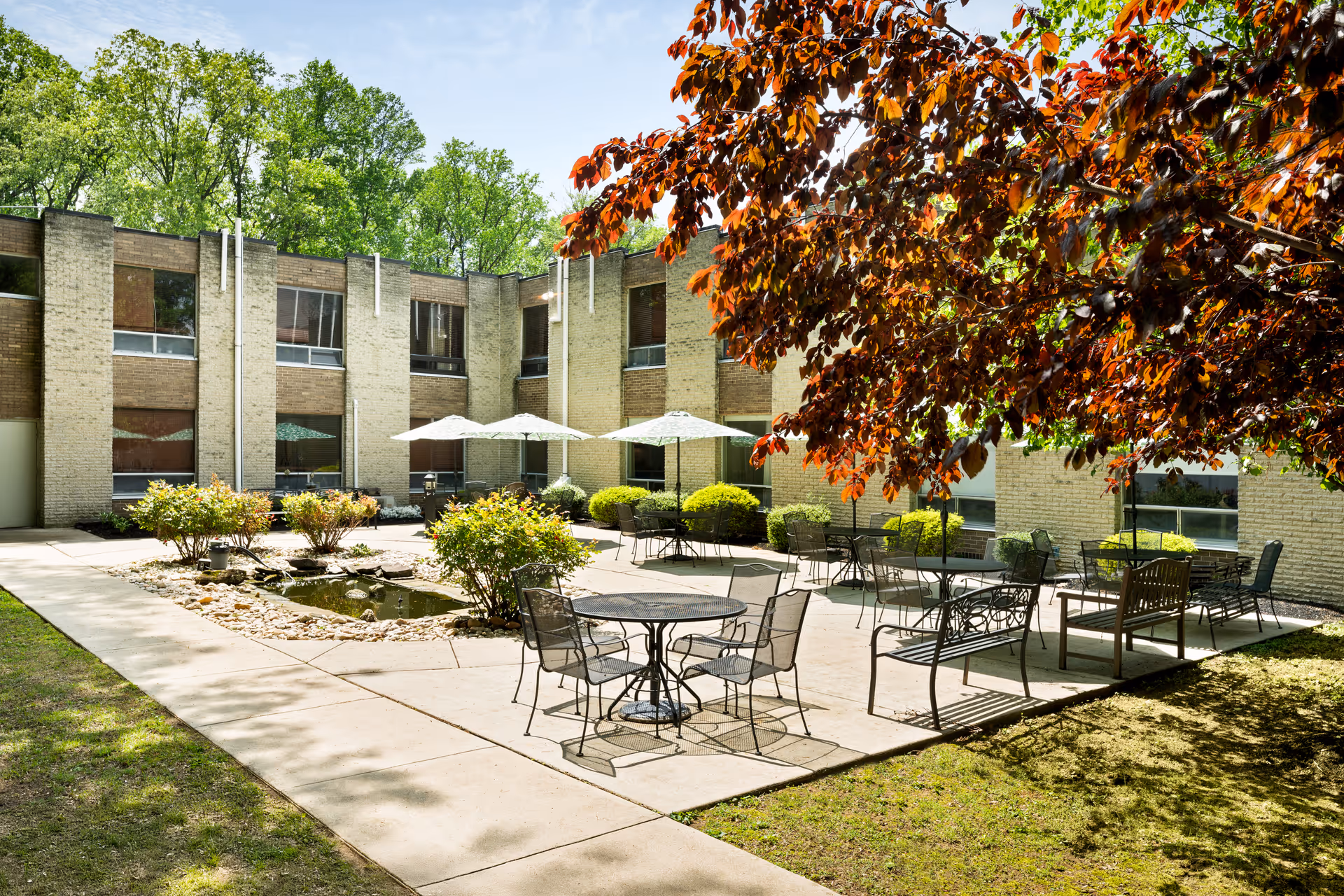 Outdoor courtyard area at Westgate Hills Rehabilitation & Nursing Center featuring metal tables and chairs, benches, umbrellas, a small pond with rocks, surrounded by a two-story brick building and trees with green and reddish leaves.