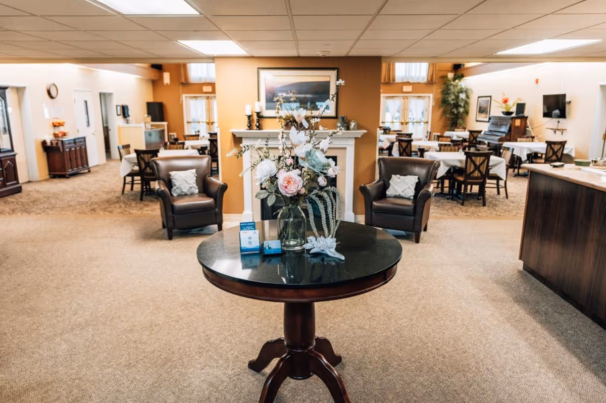 Interior view of a senior living facility common area with a round table in the foreground holding a vase of flowers and informational cards. Behind the table are two leather armchairs with pillows, a fireplace with a framed picture above it, and several dining tables with chairs in the background. The space is well-lit with ceiling lights and has a warm, inviting atmosphere.