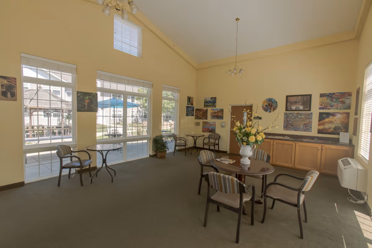 A bright common area with high ceilings and large windows letting in natural light. The room has several small round tables with chairs arranged around them. There is a vase with yellow flowers on one table. The walls are decorated with various colorful paintings and photographs. Outside the windows, a gazebo and patio area with umbrellas and seating are visible.