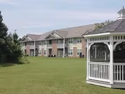 A two-story apartment building with balconies and a large grassy lawn in front. On the right side of the image, there is a white wooden gazebo with a shingled roof. Trees and shrubs are visible on the left side of the lawn.