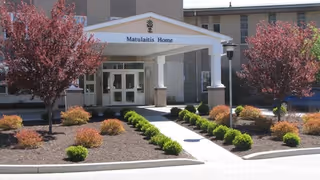 Entrance of Matulaitis Nursing Home with a covered walkway, landscaped bushes, and two red-leaved trees on either side of the path leading to the building doors.