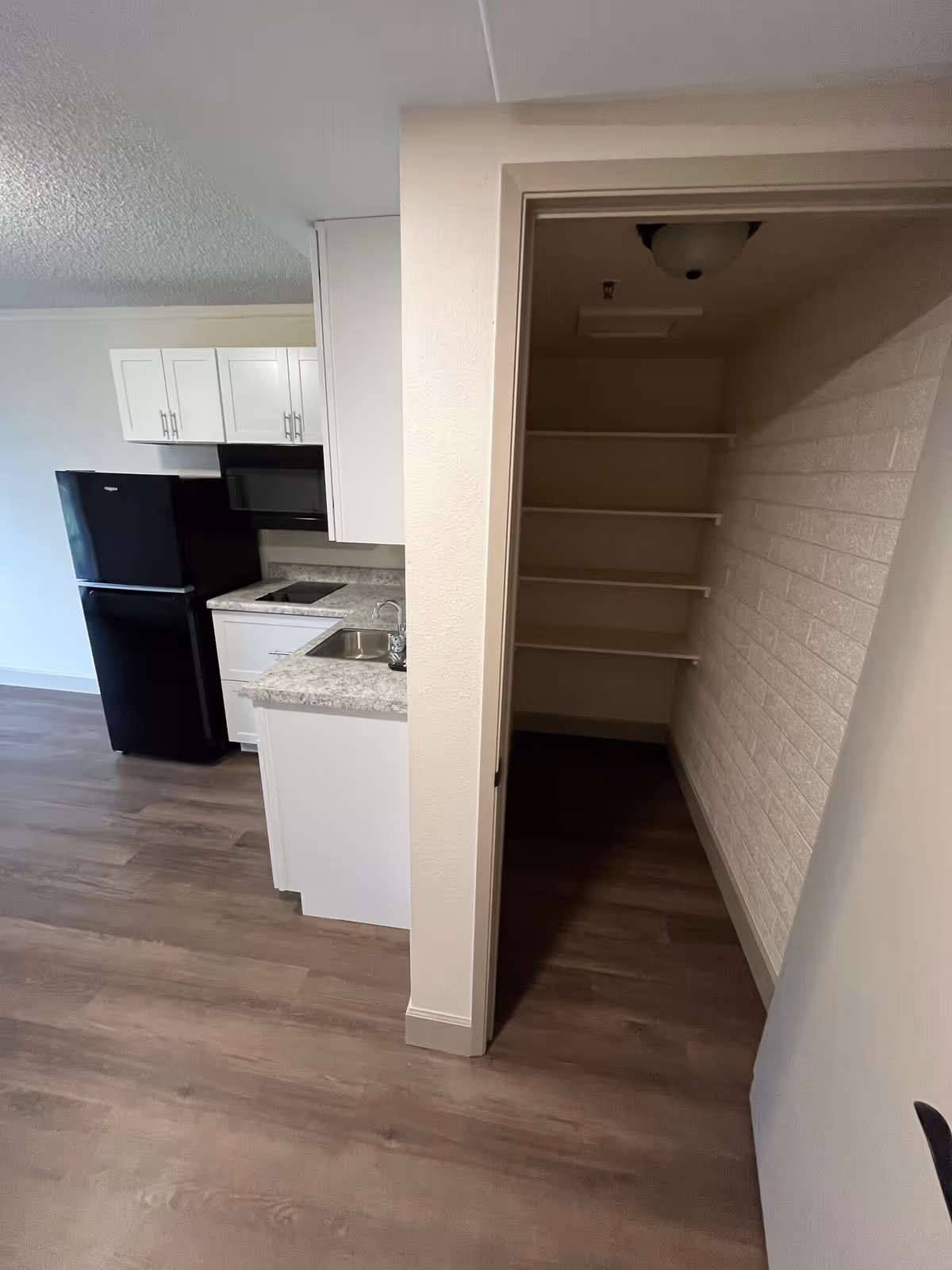 Interior view of a kitchen area with white cabinets, a black refrigerator, a microwave, a sink, and a countertop. To the right, there is an open pantry with built-in shelves and a brick-patterned wall.