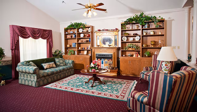 Cozy assisted-living common room with a sofa, striped armchairs, coffee table, patterned rug, and a fireplace flanked by bookshelves.