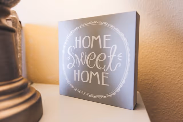 A decorative sign with the phrase 'Home Sweet Home' written in white cursive and block letters, placed on a white surface next to a lamp base against a beige textured wall.