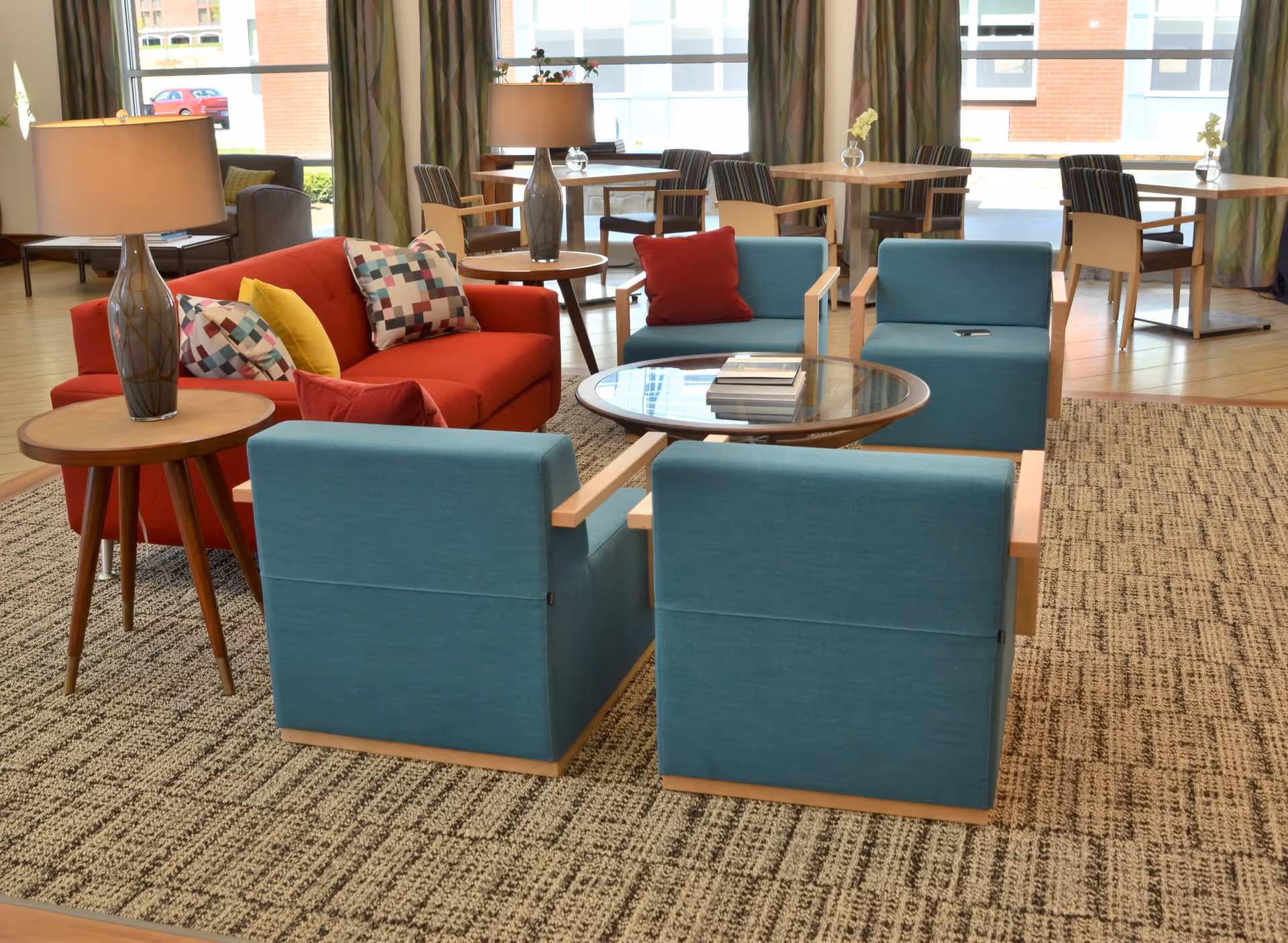 Bright common room with teal armchairs, a red sofa, round glass coffee table, side tables with lamps, and dining tables by large windows.