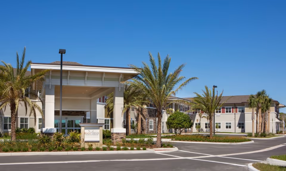 Exterior view of Beach House Assisted Living & Memory Care facility with a covered entrance, palm trees, and a clear blue sky.
