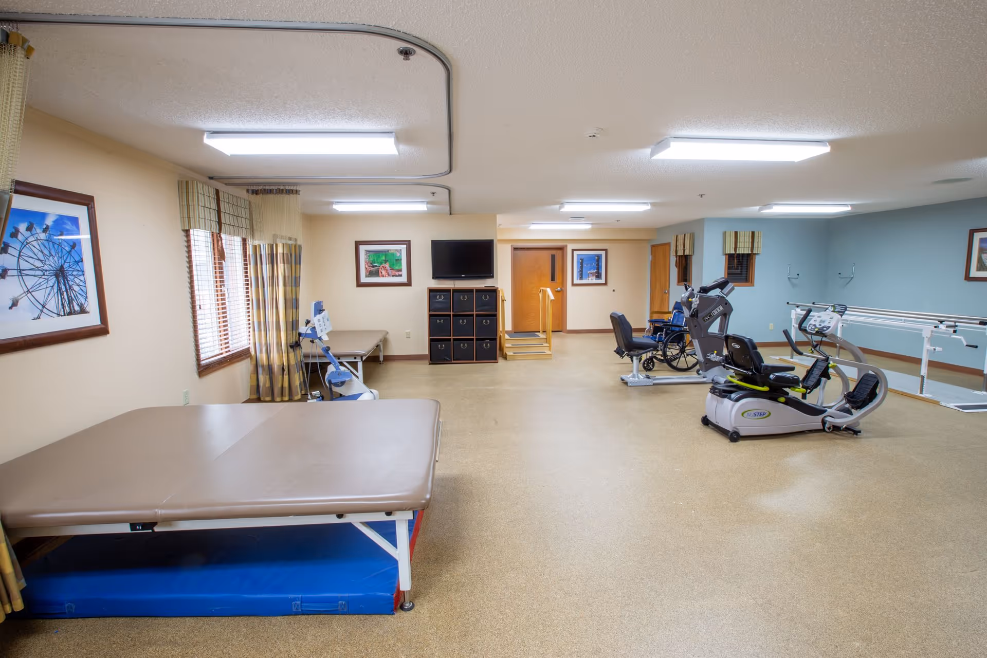 A spacious rehabilitation or physical therapy room with exercise equipment including a recumbent bike, wheelchair, parallel bars for walking support, and padded therapy tables. The room has beige walls, windows with plaid curtains, framed pictures on the walls, and bright overhead lighting.