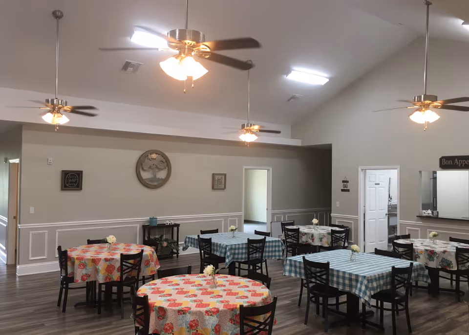 Community dining room with multiple round and rectangular tables covered in patterned tablecloths, ceiling fans, and a serving window on a neutral-colored wall.