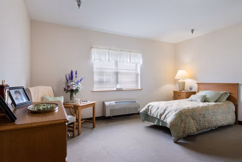 A simple furnished bedroom with a single bed, nightstand, dresser and chair beneath a window.