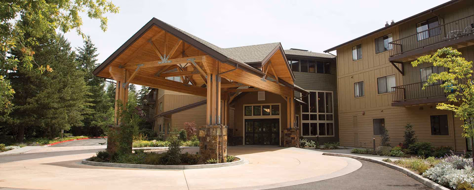 Entrance of a senior living facility with a large wooden covered driveway and surrounding greenery, including trees and shrubs. The building is multi-story with balconies and large windows.