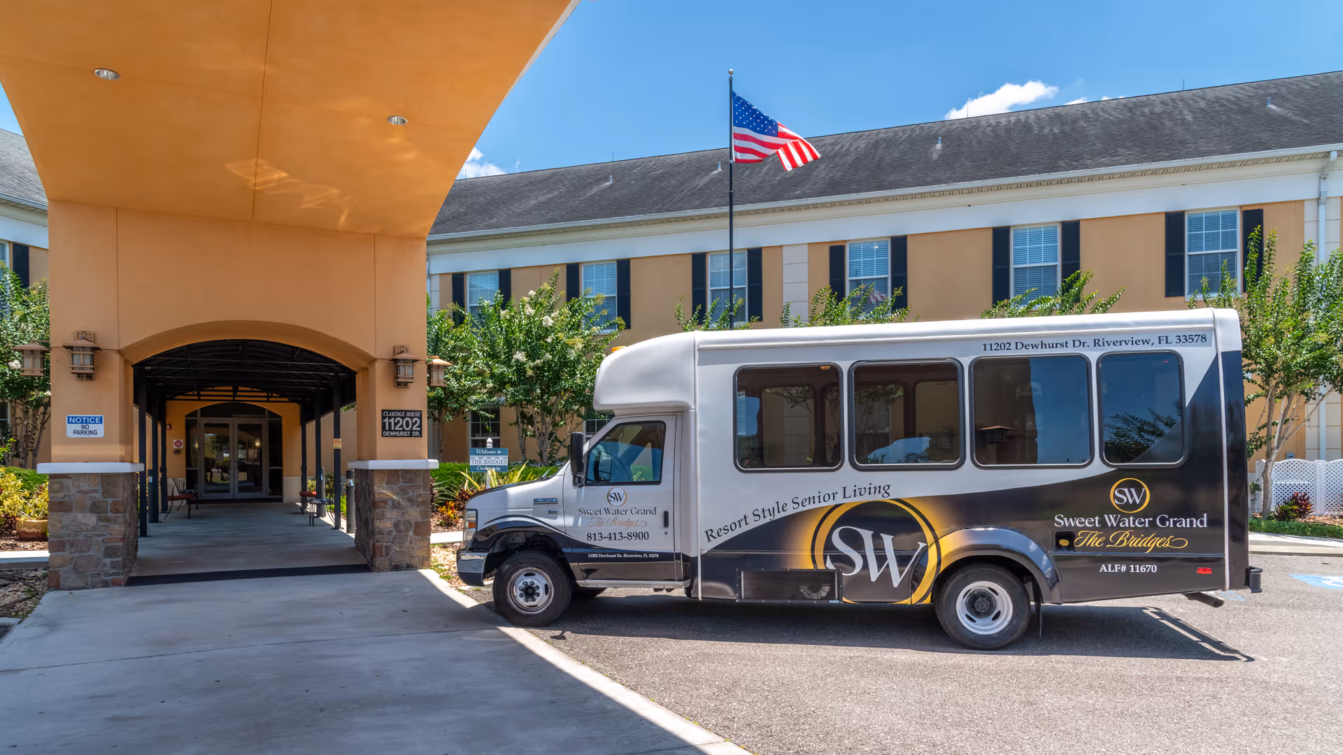 Exterior view of Sweet Water Grand The Bridges Assisted Living facility showing the entrance with a covered driveway. A shuttle bus with the facility's branding is parked in front. The building has multiple windows, an American flag on a pole, and some greenery around the entrance.