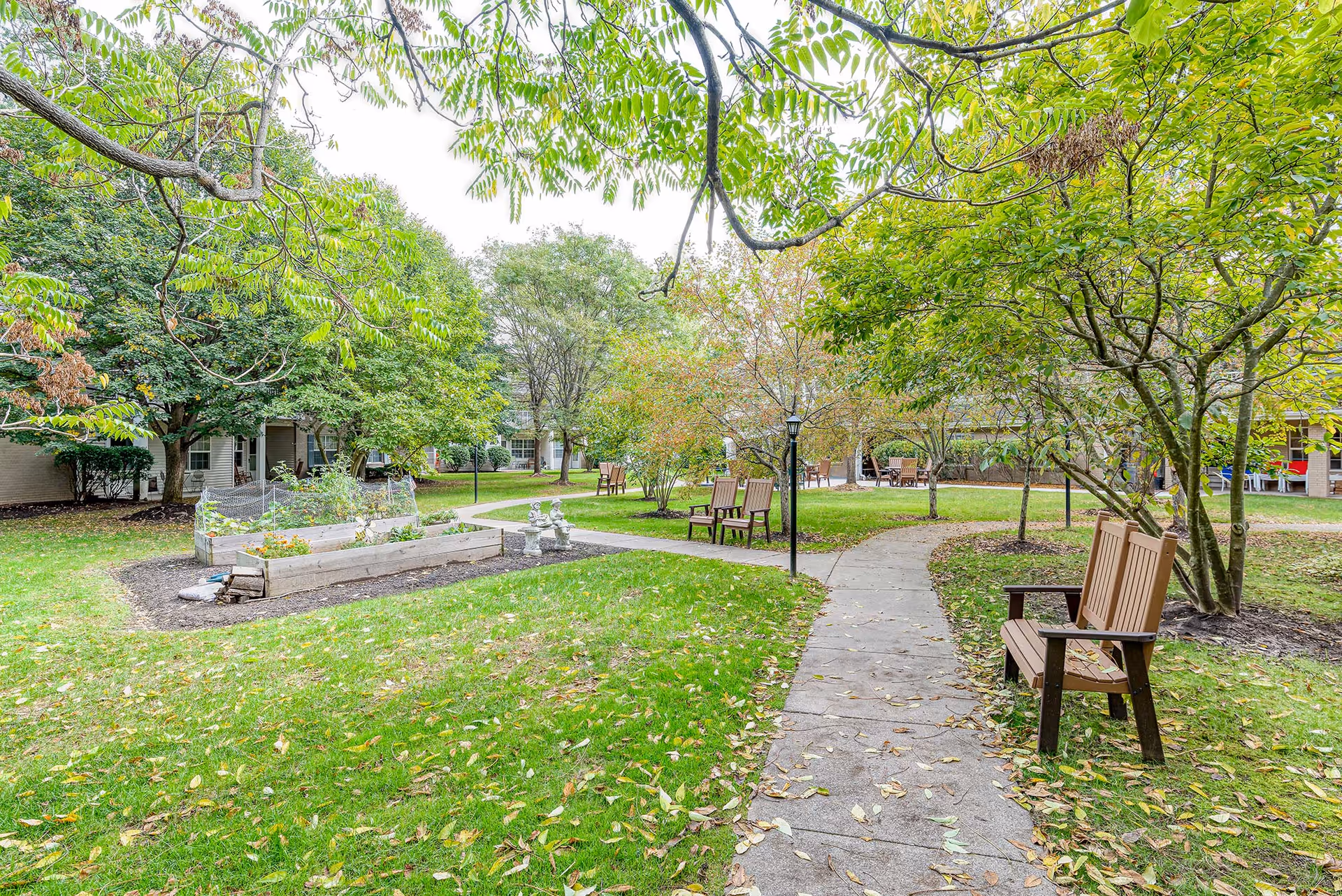 A peaceful outdoor garden area at Solstice Senior Living at Fairport featuring a paved walkway winding through green grass and trees with benches placed along the path. Raised garden beds and small statues are visible, with buildings in the background partially obscured by trees.