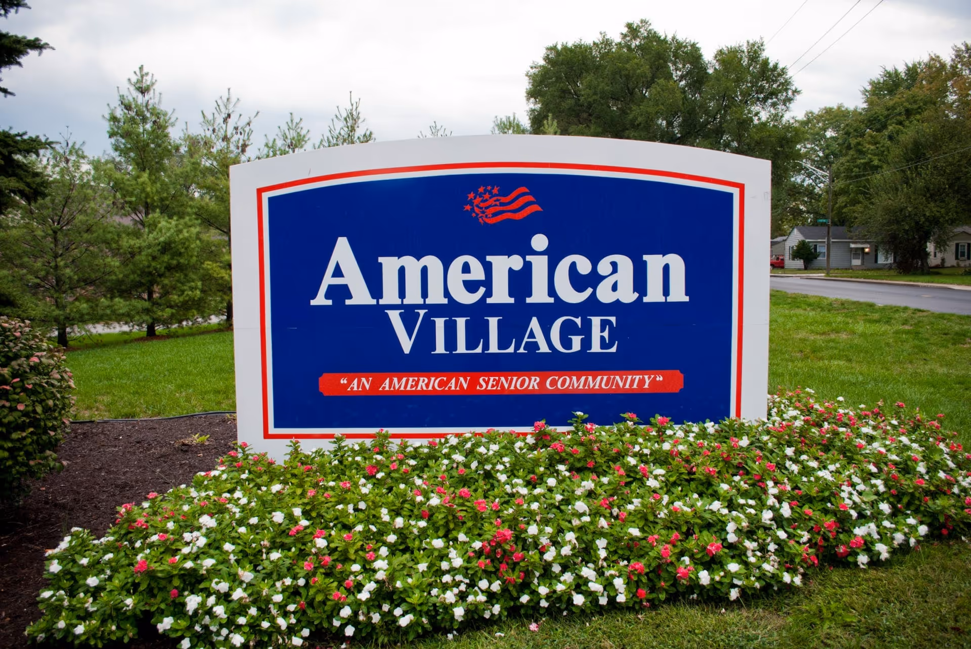 A large blue and white sign reading "American Village" sits in a flowerbed outside a senior community.