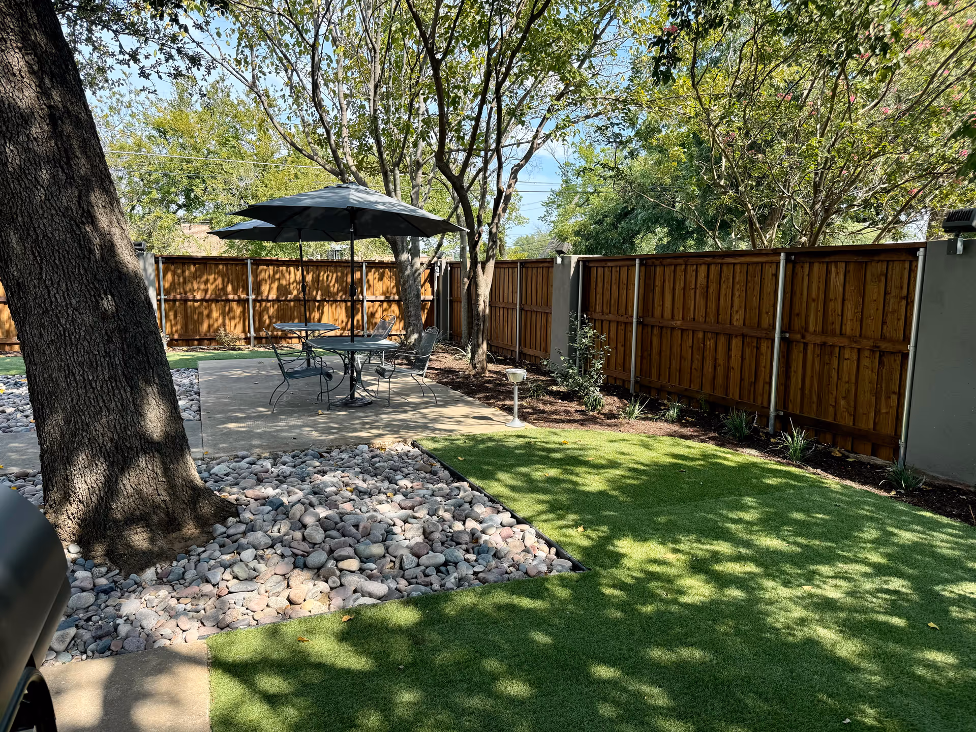 Outdoor patio area with a round metal table and four chairs under a large umbrella. The patio is surrounded by a wooden fence and shaded by several trees. There is a patch of artificial grass and a section with decorative rocks around the base of a large tree.