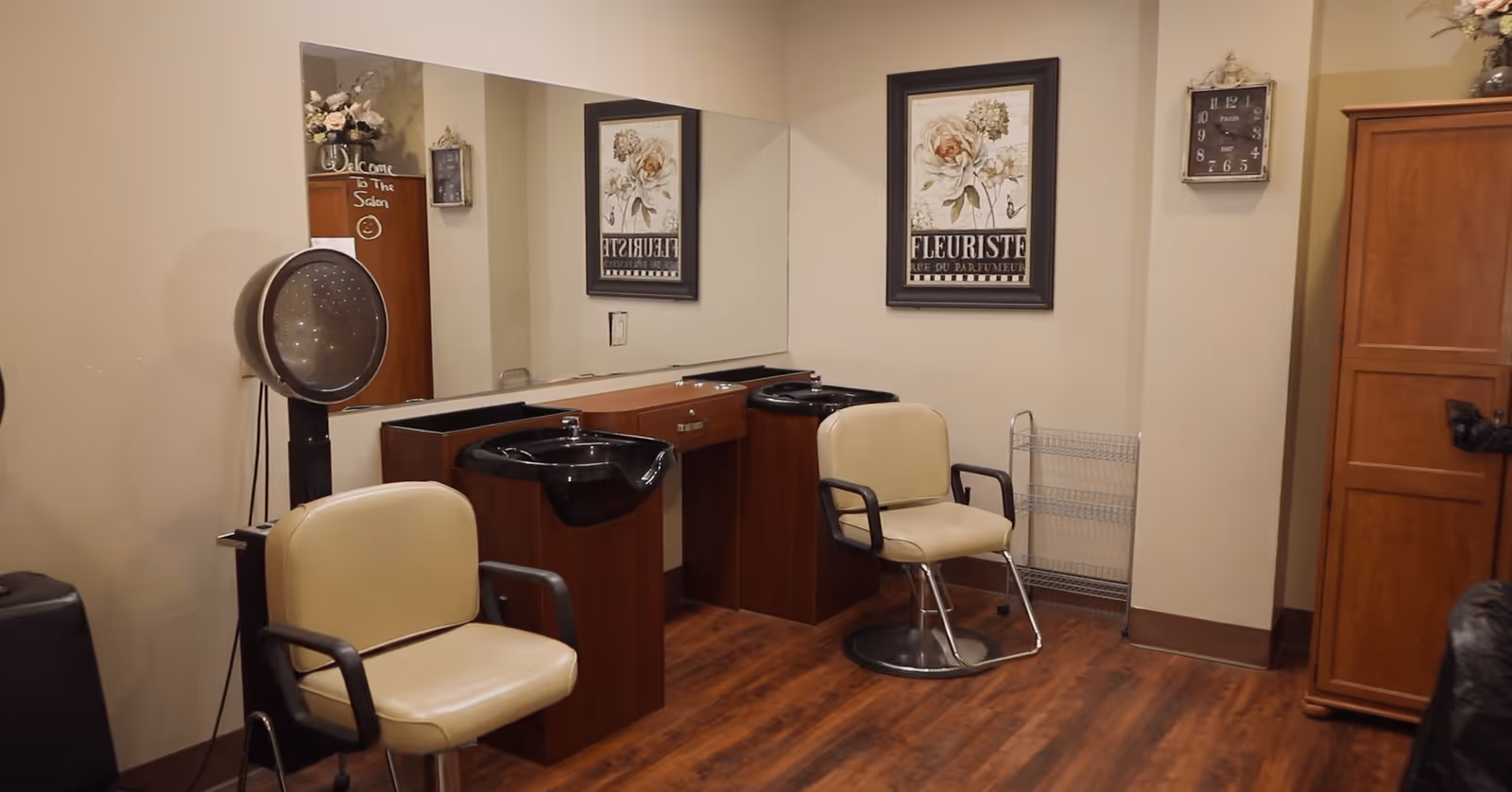 Interior of a salon area with two beige salon chairs in front of black hair washing sinks. There is a large mirror on the wall behind the sinks, a hair dryer on a stand, a wooden cabinet, a wall clock, and a framed floral picture on the wall. The floor is wooden and the walls are beige.