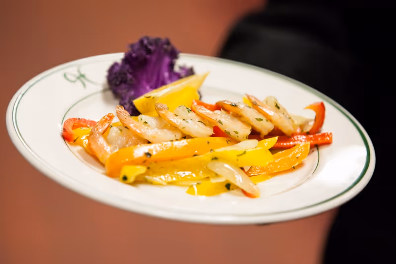 A plate of cooked shrimp served with yellow and red bell pepper strips, garnished with a lemon wedge and a piece of purple lettuce.