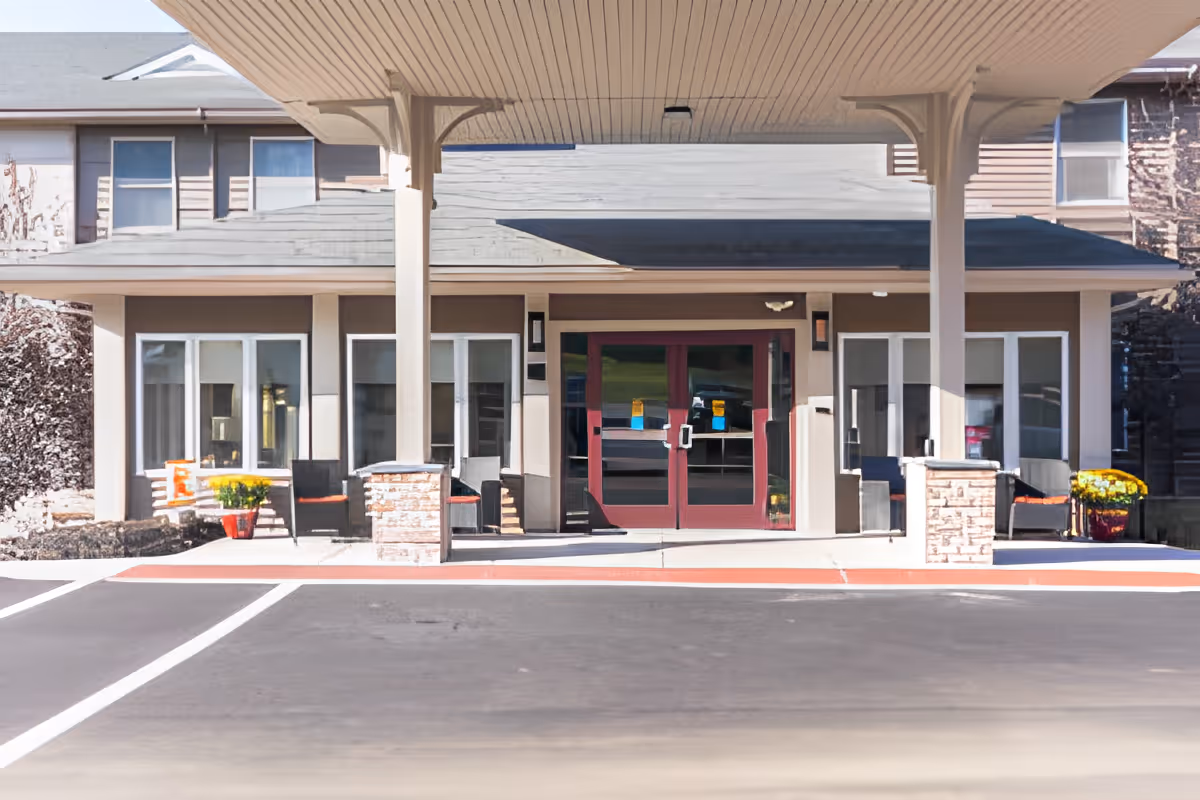 Front entrance of a senior living facility with a covered drop-off area, double glass doors, and seating areas on both sides with potted plants and chairs.