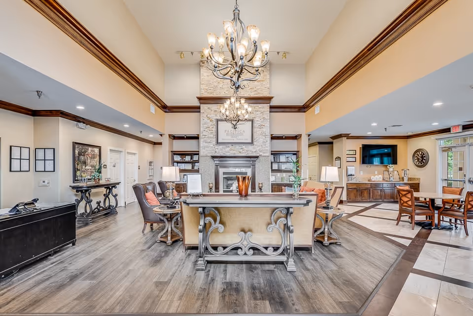 A spacious and well-lit senior living facility common area with a high ceiling, two ornate chandeliers, a stone fireplace with a framed sign above it, comfortable seating including armchairs and a sofa, wooden tables with lamps, a wall-mounted TV, and a dining area with tables and chairs. The floor is a combination of wood and tile, and the walls are painted in warm neutral tones with dark wood trim.