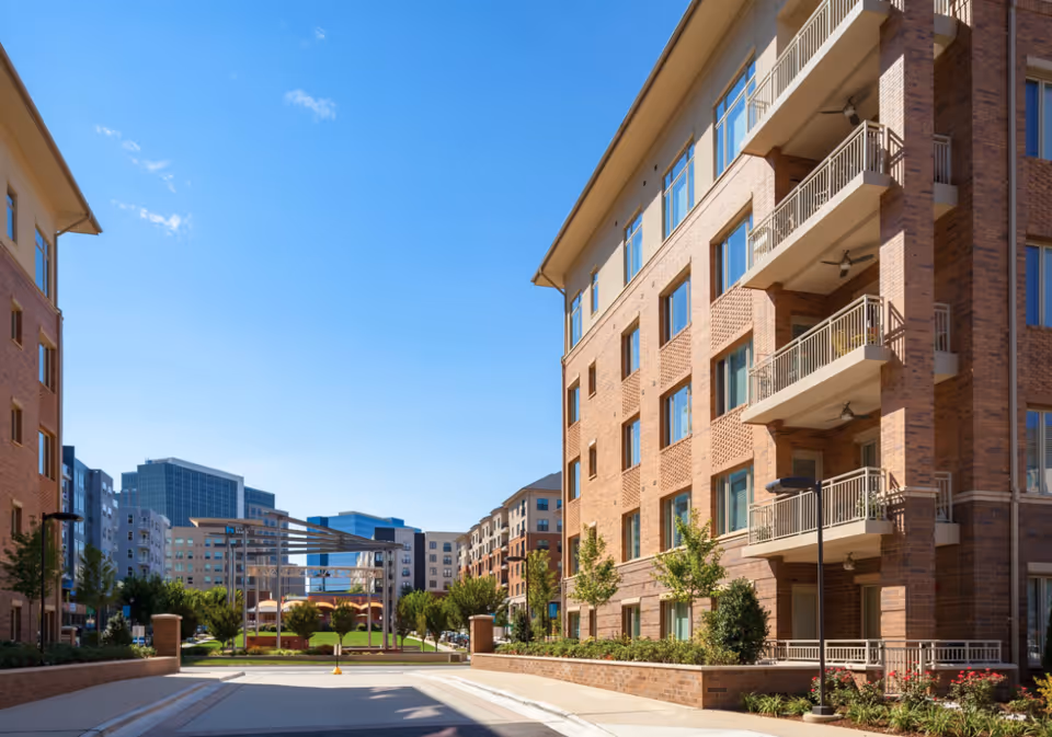 View of a modern senior living facility with multi-story brick buildings on either side, a clear blue sky, and a landscaped courtyard area with trees and greenery in the center.
